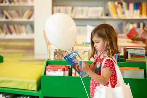 Little girl stands with a bag on her shoulder reading a book while holding a balloon