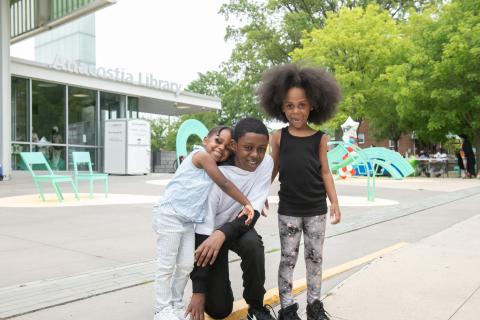 three young kids smile together in front of the Anacostia Library
