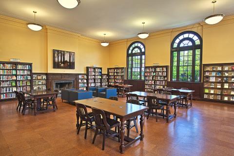 Large room with tall windows, book shelves on the outskirts of the room, and tables with chairs in the middle. 