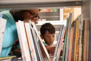 A woman and boy look through children's books on a bookshelf and the child's height.