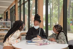 Three young girls sit at a table with a pile of books in the middle 