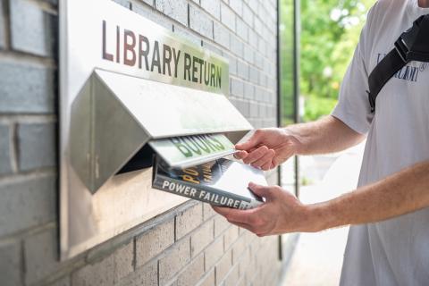 A person returns a book into the slot of an outdoor book drop at a library