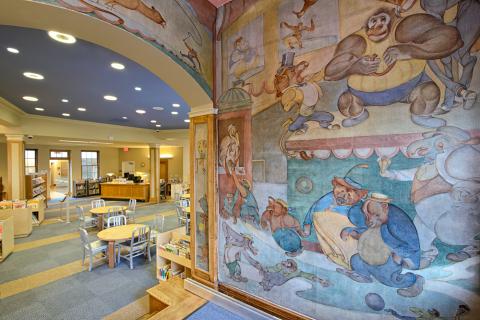 Mural of storybook characters and animals on a wall leading into a reading room with tables and chairs