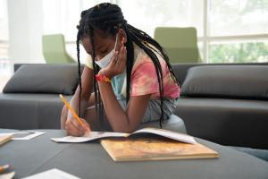 Girl in a tie dye tee shirt and shorts sits at a table writing in a workbook.