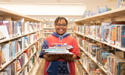 Upper elementary or middle school aged child holds a large stack of books and smiles while standing in an aisle surrounded by library shelves.