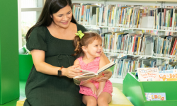 Adult and young child reading a book together on a library bench, both smiling.