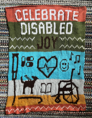 Tapestry stitched with the words Celebrate Disabled Joy with colorful stripes and icons representing assistive devices, medicine, a service dog, music notes, and a smiley face