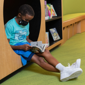 A boy reads a graphic novel sitting stretched out in a circular tunnel cutout in a library bookcase.