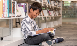 Elementary school aged child sitting cross-legged on a carpeted library floor reading a book.