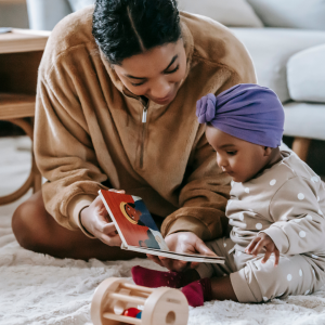On the carpet of a living room, an adult holds an open book for a sitting baby to look at, with toys laying around them.