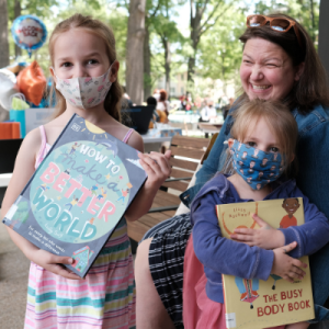 Two young children happily hold up picture books while the grown up with them smiles behind them.